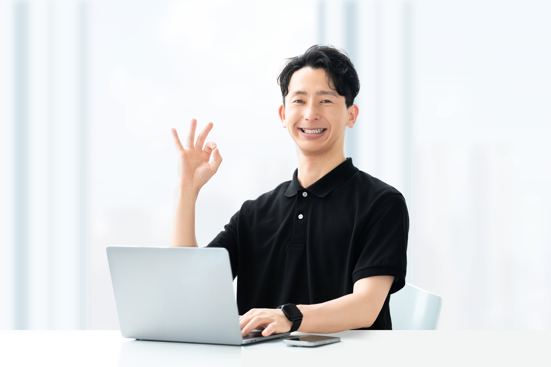 白いシャツを着た男性がノートパソコンの前に座り、満面の笑みでOKサインを見せている。