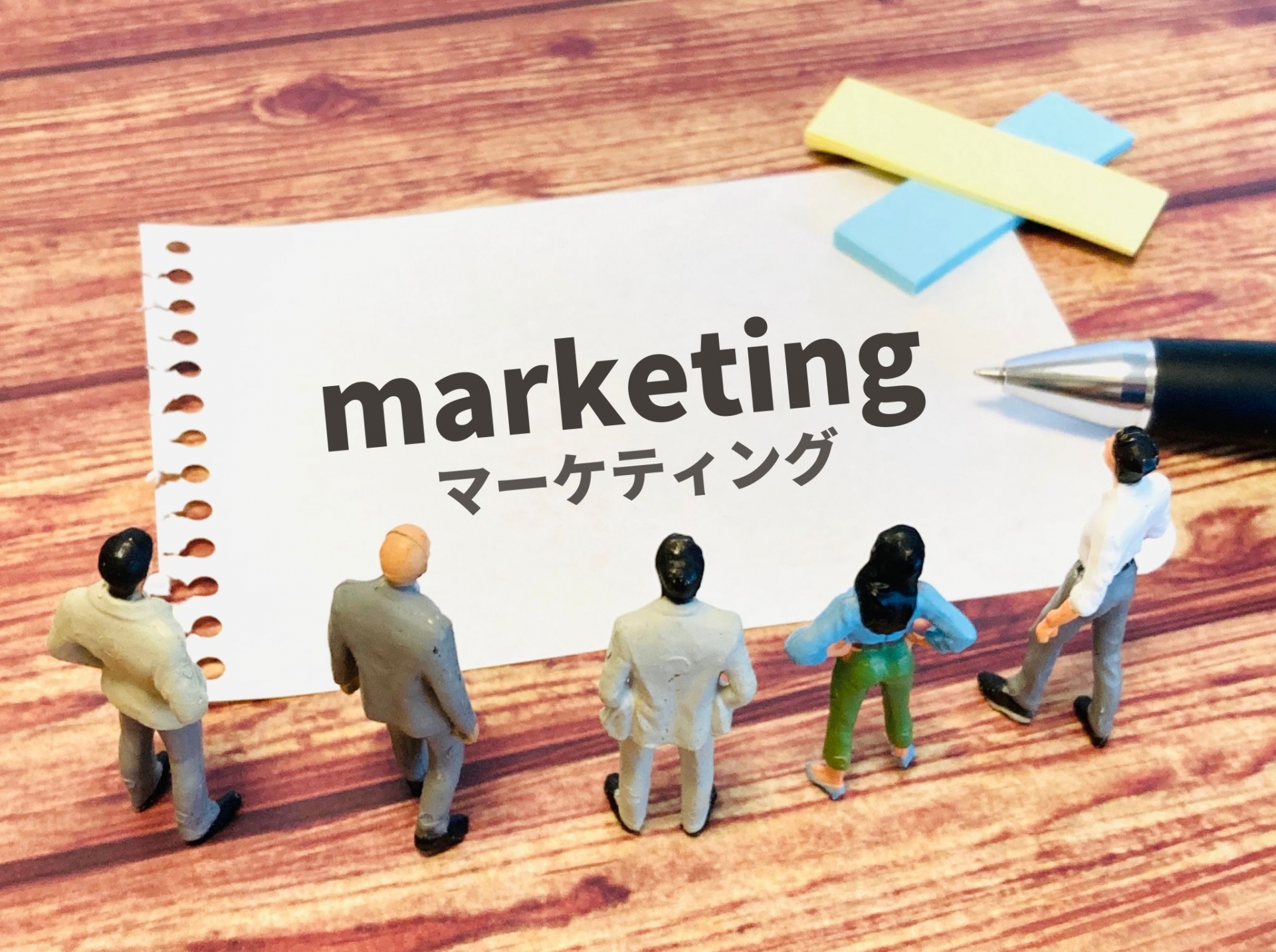 マーケティングと書かれた紙を囲むビジネスパーソンの模型